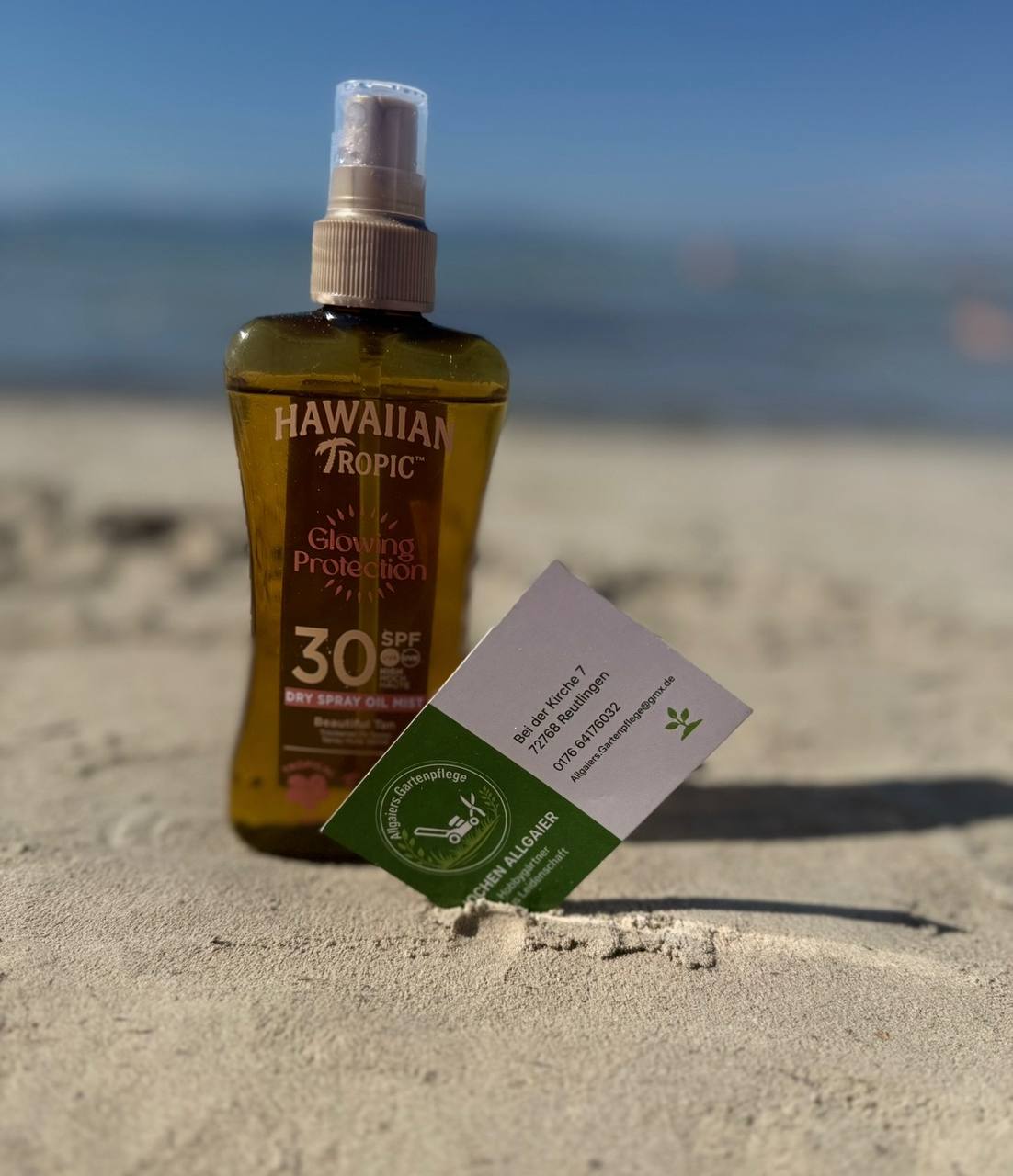 Sonnenöl und eine Visitenkarte von Allgaiers Gartenpflege im Sand am Strand